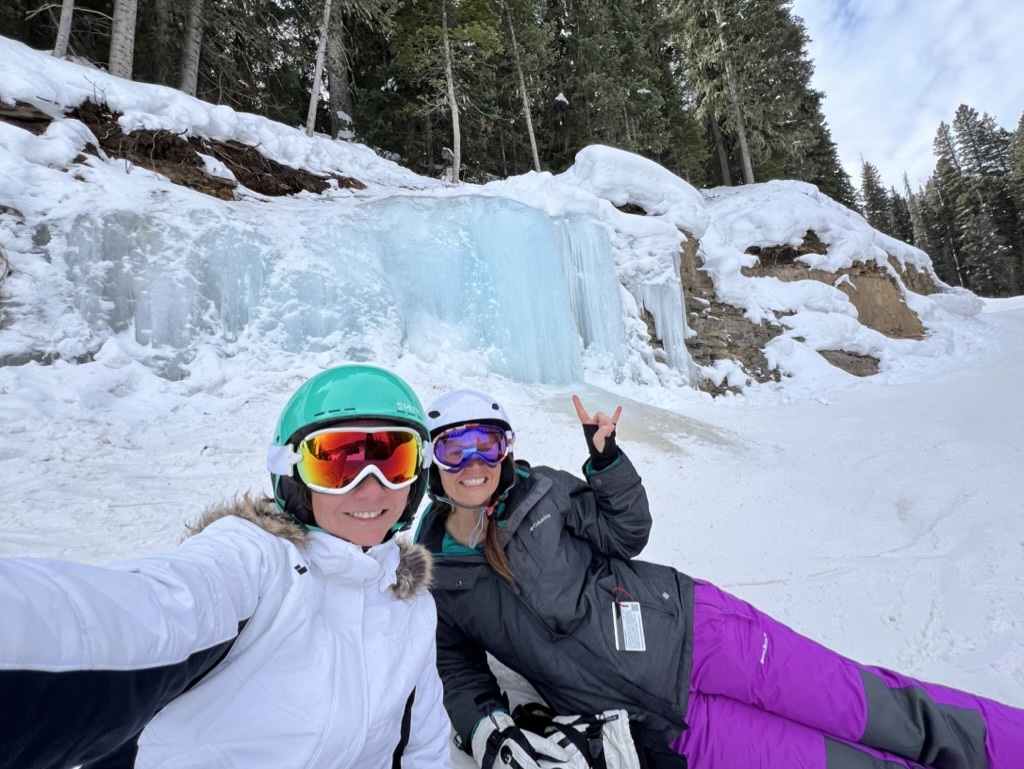 Wendy and a friend in ski helmets and goggles, smiling in front of a frozen waterfall